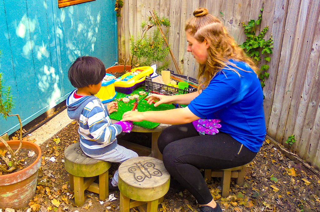 Child and Nursery Worker sat on wooden stools at a table outdoors